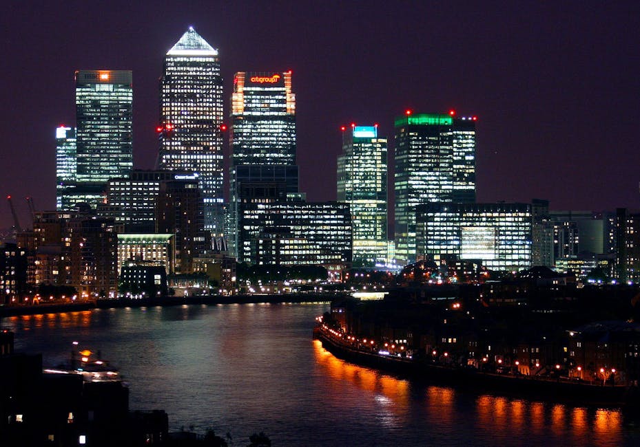 A panoramic view of Canary Wharf in London E14 during early evening, showcasing a mix of modern high-rise office towers made of glass and steel, with reflective surfaces that glisten under the soft, n