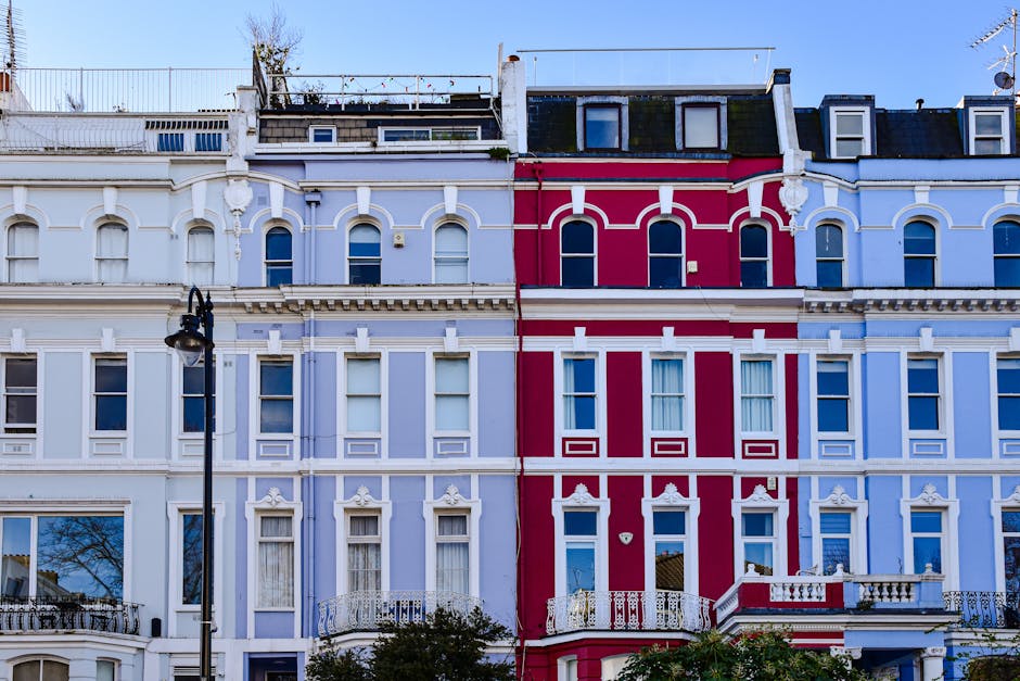 Close-up view of the exterior of a row of Victorian terraced houses in Notting Hill, London, featuring colorful facades painted in shades of blue, white, and red. The buildings have large sash windows