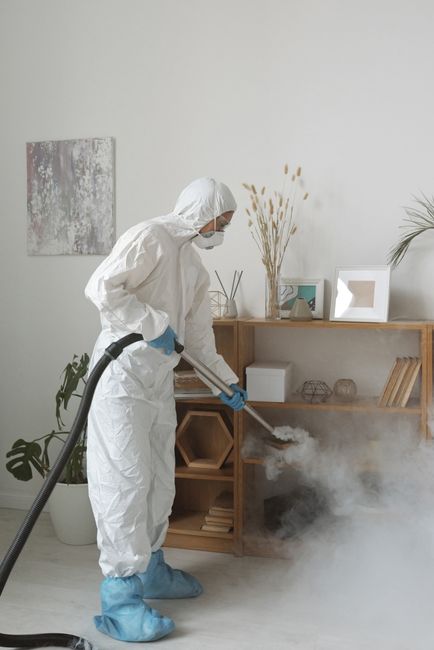A professional in full protective cleaning gear, including a white suit, gloves, mask, and shoe covers, is performing deep surface cleaning in a modern living room. The worker is using a vacuum or ste