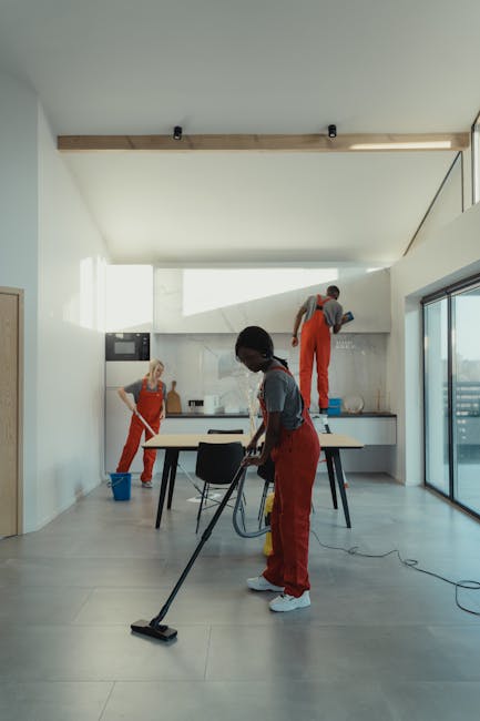 A professional in full protective cleaning gear, including a white suit, gloves, mask, and shoe covers, is performing deep surface cleaning in a modern living room. The worker is using a vacuum or ste